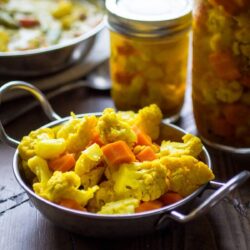 A jar and a bowl with Curried Carrot and Cauliflower Pickles on a table.