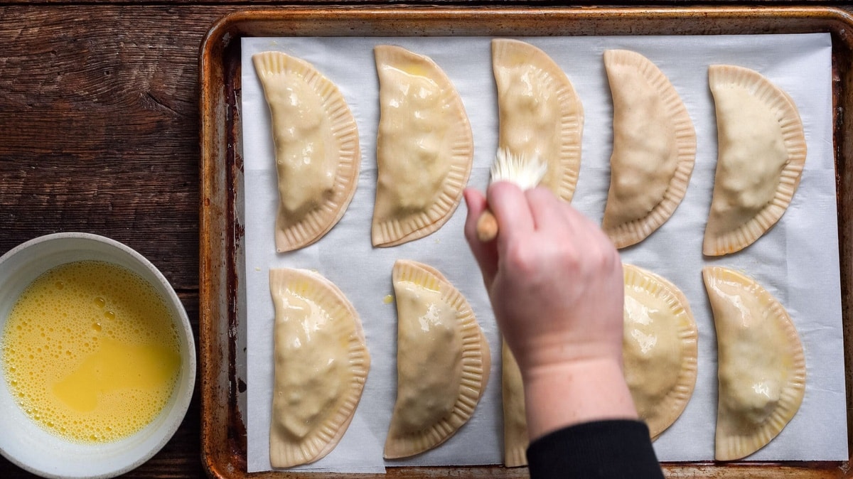 Beef empanadas being brushed with egg wash while on a cookie sheet.